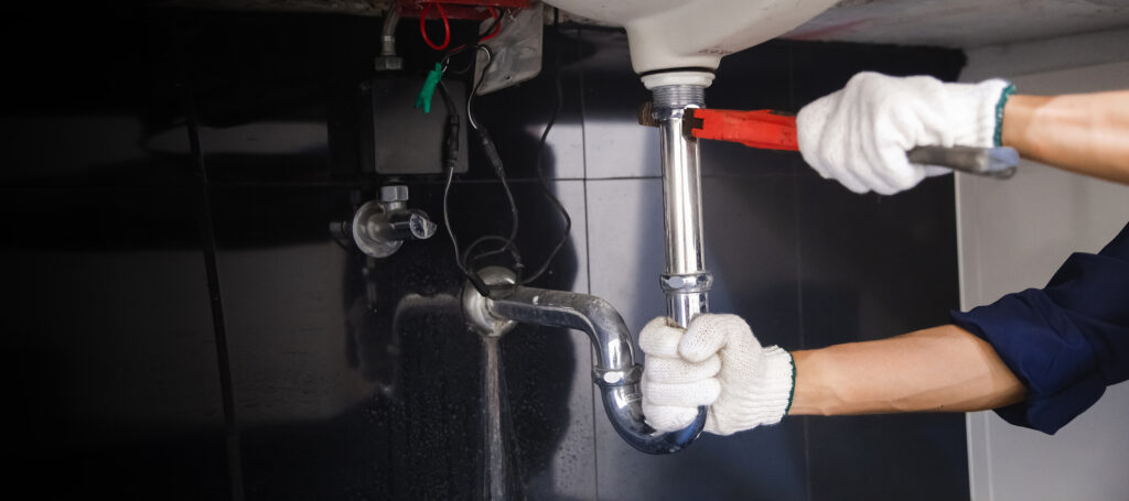 Gloved plumber tightening a chrome drain pipe beneath a sink.