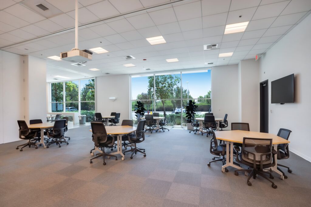 Bright training classroom with table groups, black chairs and windows overlooking trees.