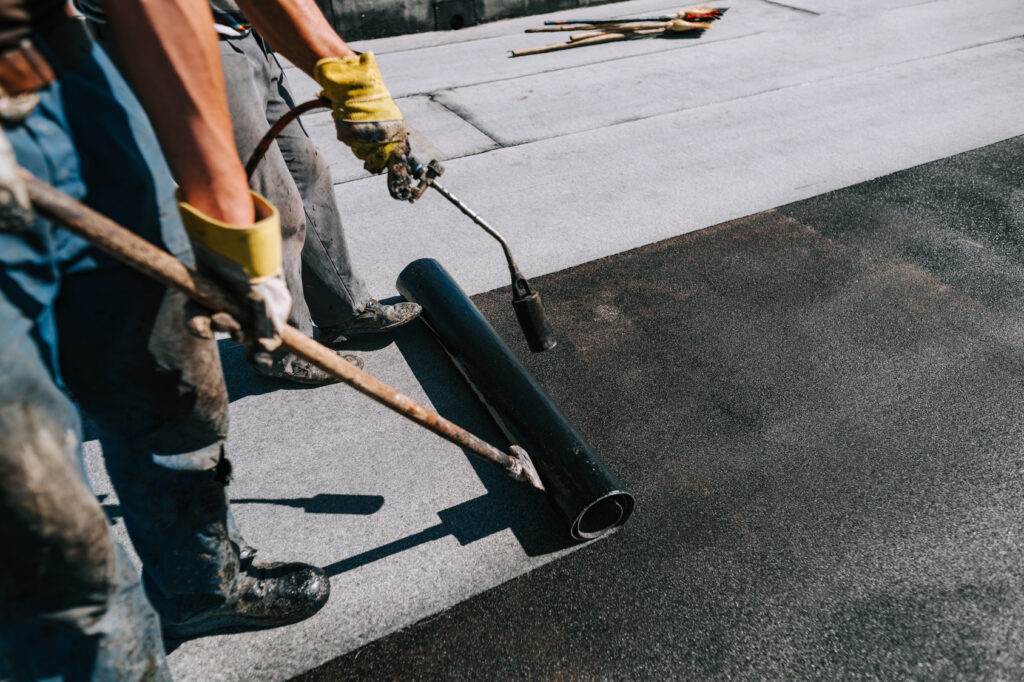 Gloved roofing worker torch-applying a roll of bituminous membrane.