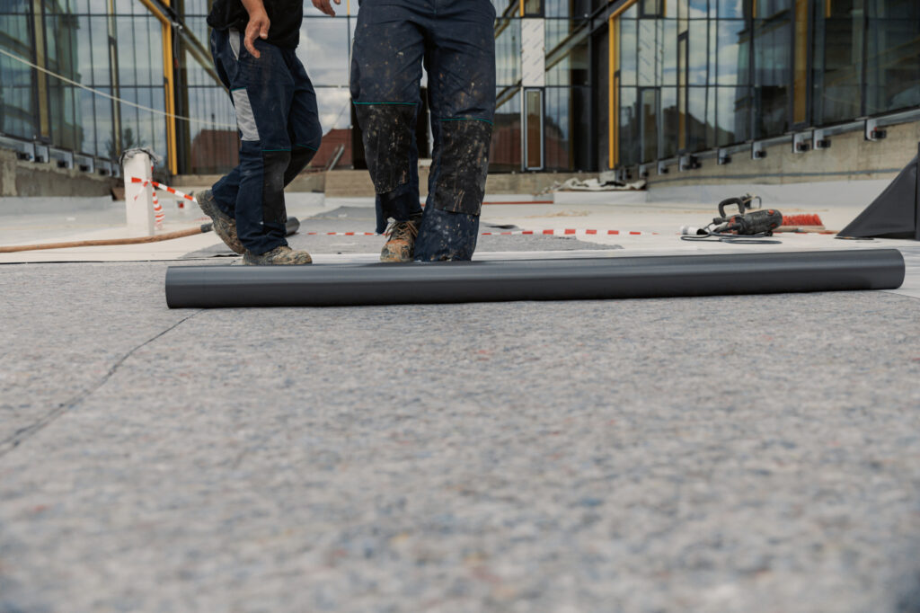 Roofing membrane roll beside workers' boots on a flat rooftop.