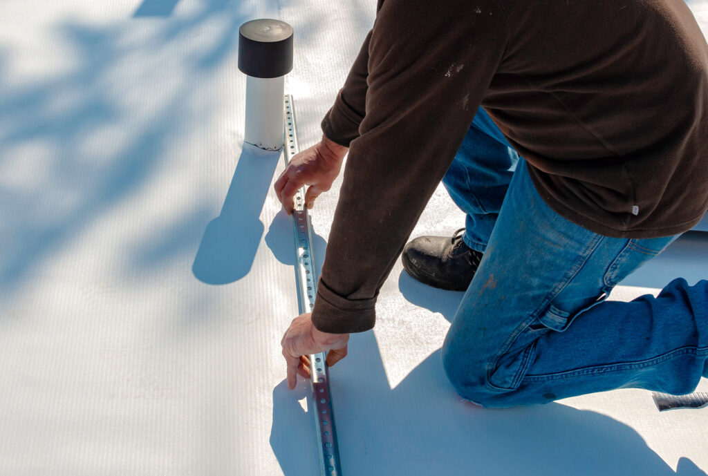 Worker positioning a metal strip on a light-colored roof membrane beside a pipe penetration.