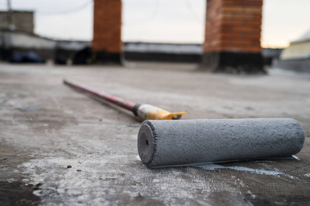 Paint roller resting on a flat roof surface with roof penetrations in the background.